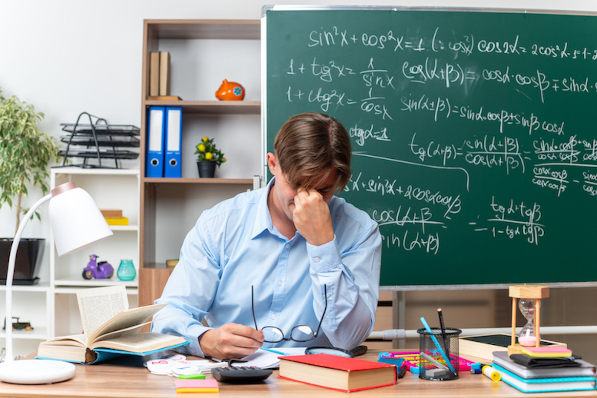 young-male-teacher-wearing-glasses-tired-overworked-touching-his-nose-closed-eyes-sitting-school-desk-with-books-notes-front-blackboard-classroom.jpg